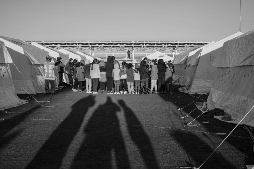 Black and white photo of a group gathered between tents in a Turkish refugee camp.